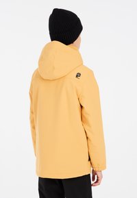Veste imperméable jaune avec capuche, poignets élastiques, détail du logo au dos, associée à un bonnet en tricot noir. Fini lisse et mat.