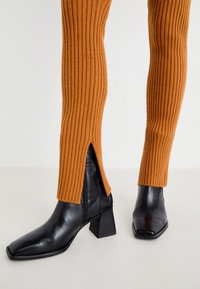 Zwarte leren enkellaars met vierkante neus en stevige hak, gecombineerd met oranje geribbelde broek met slits bij de enkel.
