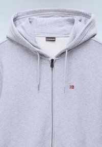 Sweat à capuche gris clair avec fermeture éclair, cordons, petit logo drapeau norvégien sur la poitrine, et étiquette de marque à l'intérieur du col.