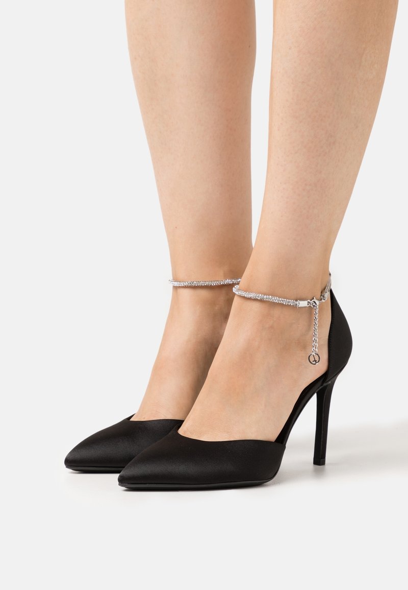 Escarpins noirs à bout pointu avec fines brides de cheville ornées de strass sur jambes nues, debout sur un fond clair.