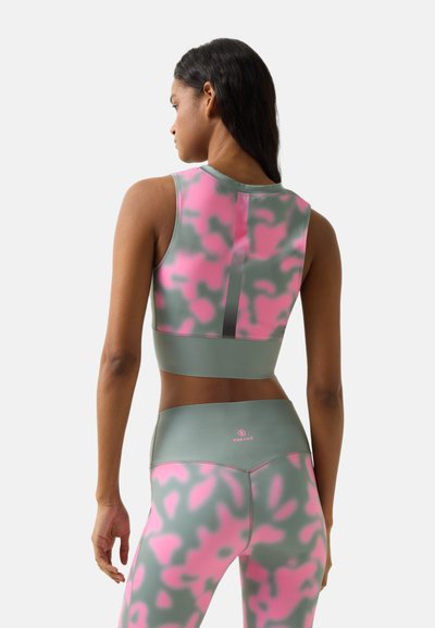 Femme portant un haut court sans manches à motif camouflage vert et rose ainsi que des leggings assortis, debout dos à la caméra.