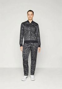 Μαύρο αθλητικό κοστούμι με υφή σε σχέδιο ζώου, φερμουάρ στο μπουφάν και tapered παντελόνι, συνδυασμένο με λευκά αθλητικά παπούτσια.