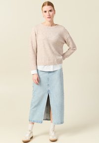 Maglione a maglia beige chiaro con orlo a costine e bottoni sulle spalle, indossato sopra una gonna maxi in denim azzurro chiaro con spacco frontale. Sneakers bianche.