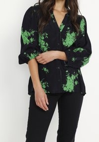 Chemise boutonnée bleu marine foncé avec imprimé floral vert. Caractéristiques : manches bouffantes, coupe décontractée et texture douce. Porte un pantalon noir.
