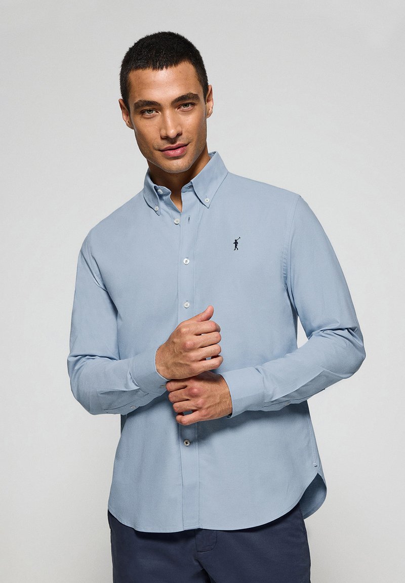 Light blue button-up shirt with long sleeves, tailored fit, and a small black embroidered logo on the chest. Smooth fabric, crisp collar.