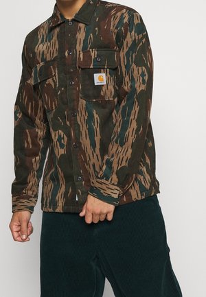 Chemise en velours côtelé avec un motif camouflage, présentant des rayures marron, vert et beige. Deux poches poitrines et étiquette de marque sur la poche gauche.