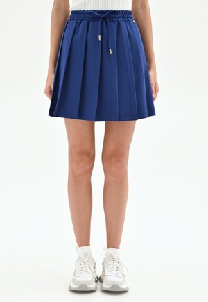 Femme portant une jupe plissée bleue à taille à cordon, associée à des baskets blanches et des chaussettes, debout devant un fond uni.