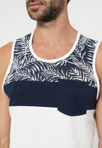 Homme portant un débardeur sans manches avec un motif de feuilles de palmier blanc, bleu marine et blanc, avec une petite poche poitrine bleu marine.