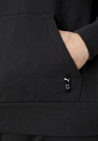 Sudadera negra con capucha que presenta un bolsillo tipo canguro y puños acanalados. Muestra una etiqueta con el logo de Puma en el bolsillo para la identificación de la marca. Textura suave.