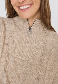 Beżowy sweter na zamek błyskawiczny z dzianiny, z prążkowanym kołnierzem, wzorami warkoczowymi i teksturowanym materiałem. Akcentujący detal z suwakiem w kolorze srebrnym.