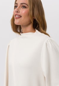 Crème-kleurige blouse met pofmouwen en een hoge hals. Gemaakt van zachte stof, met een gladde textuur en een minimalistisch ontwerp.