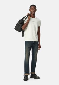 Camiseta blanca con el diseño de texto "EN ESCENA", jeans azul oscuro, mochila negra y zapatos negros. El modelo está de pie con la mochila sobre el hombro izquierdo.
