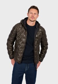 Man wearing dark quilted jacket with hood, black sweater, and blue jeans, standing against plain light gray background.