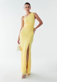 Asymmetrisches gelbes Kleid aus strukturiertem Stoff mit hohem Schlitz. Kombiniert mit weißen Riemchenschuhen und einer kleinen weißen Handtasche. Elegantes Design.