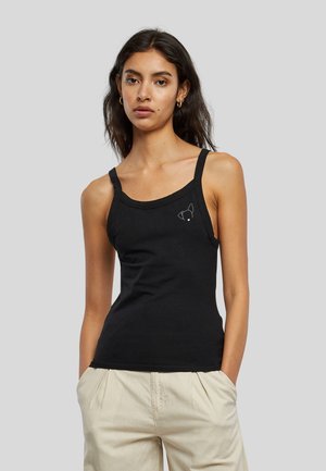 Femme aux longs cheveux foncés portant un débardeur noir avec un petit logo de chien et un pantalon beige clair, debout les mains dans les poches.