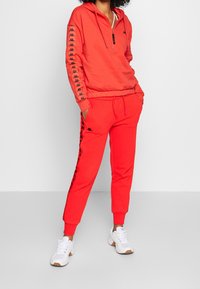 Person wearing red Kappa tracksuit with black logo stripes on sleeves and pants, paired with white sneakers, standing with hands in pockets.