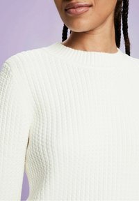 Maglione bianco a coste con un collo alto, superficie testurizzata e design aderente. Focus sui dettagli delle spalle e del collo.