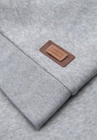 Felpa grigia con una texture morbida, caratterizzata da una patch rettangolare in pelle marrone con testo impresso e borchie argentate.
