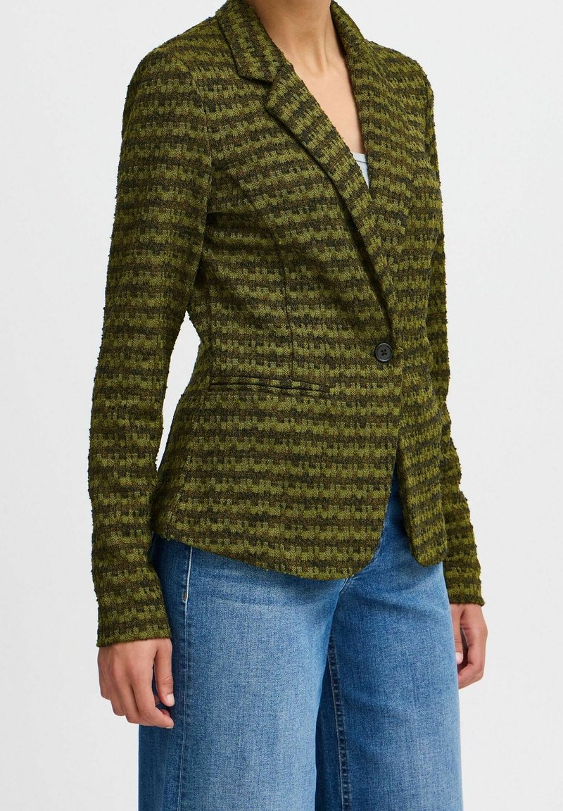 Femme portant un blazer pied-de-poule texturé vert et noir à un bouton sur un top blanc et un jean bleu.