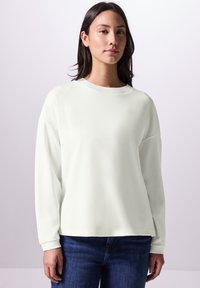 Losse, crèmekleurige sweatshirt met geribbelde halslijn en lange mouwen, gecombineerd met blauwe jeans. Zachte textuur, minimalistisch ontwerp.