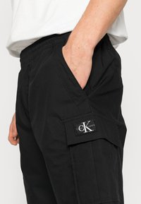 Černé cargo kalhoty s elastickým pasem, bočními kapsami a nášivkou loga Calvin Klein na levém stehně. Bavlněný materiál, klasický střih.