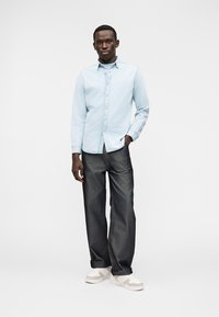 Camisa azul-clara com botões e colarinho, combinada com jeans escuros e ténis brancos com detalhes bege. Fundo liso.