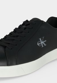 Baskets Calvin Klein noires avec lacets noirs et semelle blanche, montrant le logo latéral et la tige en cuir lisse en gros plan.