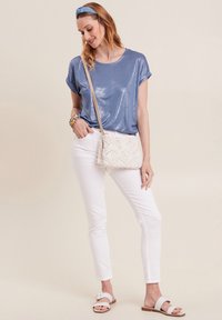 Lichtblauw, glanzend T-shirt met opgerolde korte mouwen, gecombineerd met witte, aansluitende broek en witte sandalen. Inclusief een geweven schoudertas.