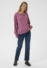 Mujer que lleva un suéter largo de mangas de pliegue color malva, jeans oscuros azules y loafers gruesos negros, de pie con una mano en el bolsillo.