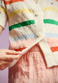 En stickad cardigan med korta ärmar, prydd med flerfärgade ränder i orange, gult, grönt och blått på en krämfärgad bakgrund. Vita knappar.