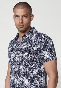 Kratke rokavov, srajca na gumbe v mornarsko modri barvi z belim tropičnim listnim vzorcem, s klasičnim ovratnikom in lesenimi gumbi.