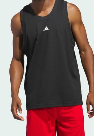 Homme portant un débardeur de sport Adidas noir et un short de sport rouge, debout devant un fond clair.