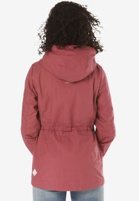 Rote, leichte Jacke mit Kapuze, elastischem Bund und einem Logo auf der Rückseite. Verfügt über eine glatte Textur und ein kleines weißes Markenzeichen auf der linken unteren Seite.