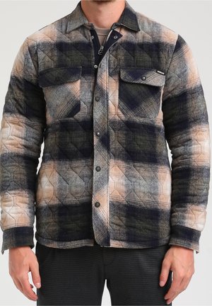 Mann trägt eine gesteppte karierte Jacke in Beige-, Schwarz- und Grautönen mit geknöpften Brusttaschen und Druckknöpfen vorne.