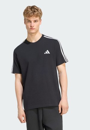 Ung mand iført en sort Adidas t-shirt med hvide striber på skuldrene og logo på brystet, stående mod en ensfarvet baggrund.