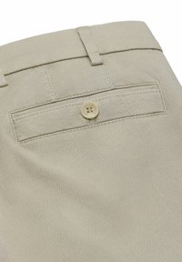 Beige rugzakzak van een broek met structuur, voorzien van een enkele knoopsluiting en riemlussen boven de tailleband.