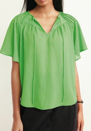 Femme portant un chemisier texturé vert clair à manches courtes, col en V et coupe ample, associé à une jupe noire.