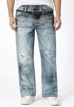 Uomo che indossa jeans blu chiaro slavati e consumati con chiavi decorative e borchie, abbinati a sneaker bianche e una camicia bianca.