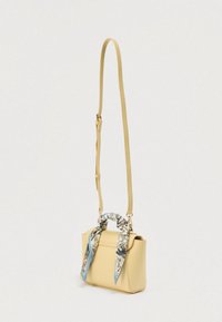 Petit sac bandoulière beige avec bandoulière réglable et une écharpe à motif floral nouée autour de la poignée supérieure sur un fond uni.