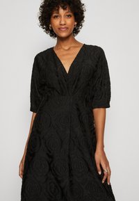 Vestido negro con un diseño floral texturizado, que cuenta con un escote en V profundo y mangas cortas abullonadas. La tela tiene un ligero brillo.
