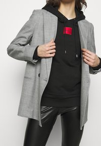 Osoba poprawiająca szary żakiet na czarnym hoodie z czerwonym logo, ubrana w czarne skórzane spodnie na jednolitym jasnym tle.