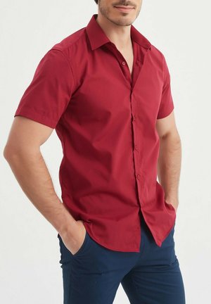Chemise rouge à manches courtes, boutonnée, en tissu léger, avec un col classique et une texture lisse, portée avec un pantalon bleu marine.