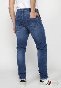 Jeans de mezclilla azul de corte ajustado con dos bolsillos traseros, detalles de costura y una textura suave, combinados con zapatillas blancas y grises.