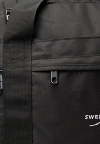 Black cargo duffel bag with textured fabric, adjustable strap, zip closure, and white logo detail. Features reinforced stitching and hardware accents.