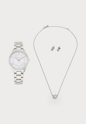 Reloj de pulsera Michael Kors en tono plateado con esfera blanca, collar de cadena plateada con colgante redondo y pendientes a juego sobre fondo blanco.