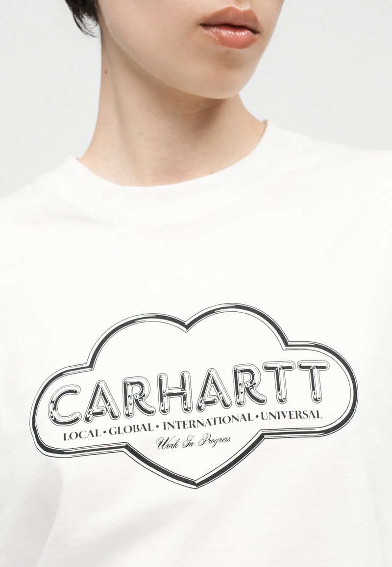 Person trägt ein weißes Hemd mit einem schwarzen, herzförmigen Logo mit der Aufschrift "CARHARTT" und den Worten "Local Global International Universal".