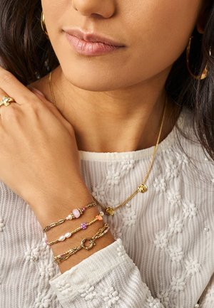 Woman wearing white textured top with gold hoop earrings, layered gold necklace, and multiple delicate gold and beaded bracelets on left wrist.