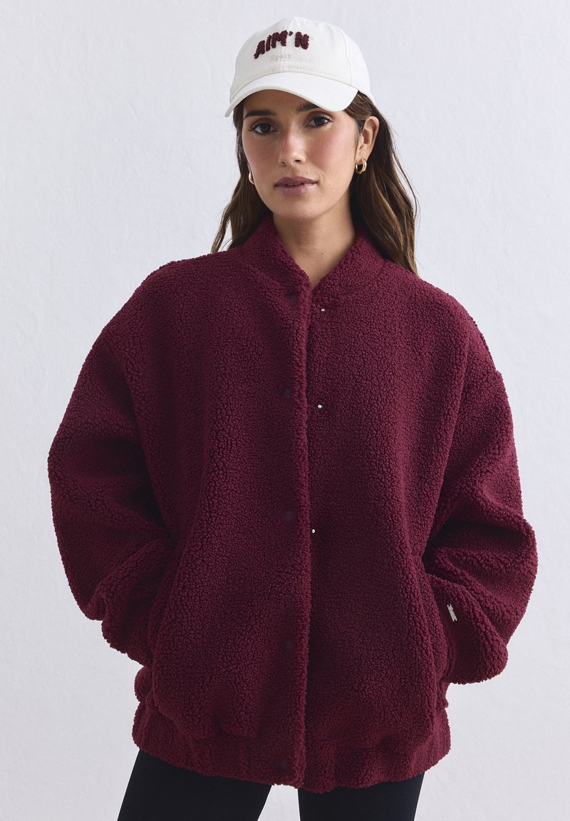 Γυναίκα που φοράει μπορντό fleece μπουφάν με κουμπιά και λευκό καπέλο μπέιζμπολ στέκεται μπροστά σε απλό ανοιχτόχρωμο τοίχο.