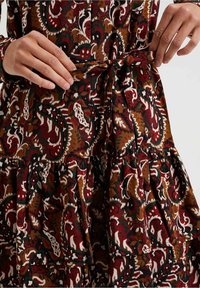 L'abito presenta un motivo paisley in tonalità di rosso, verde e marrone. Realizzato in un tessuto testurizzato con una vita annodata e un design a gonna a più strati.