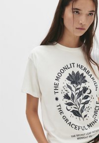 Ung kvinde med langt hår iført en hvid t-shirt med et sort blomsterdesign og teksten "The Moonlit Herbarium Graceful Min Circle."